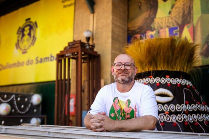 Historiador Luiz Antonio Simas, de óculos e camiseta branca, apoiado em uma mureta diante de fachada amarela de escola de samba