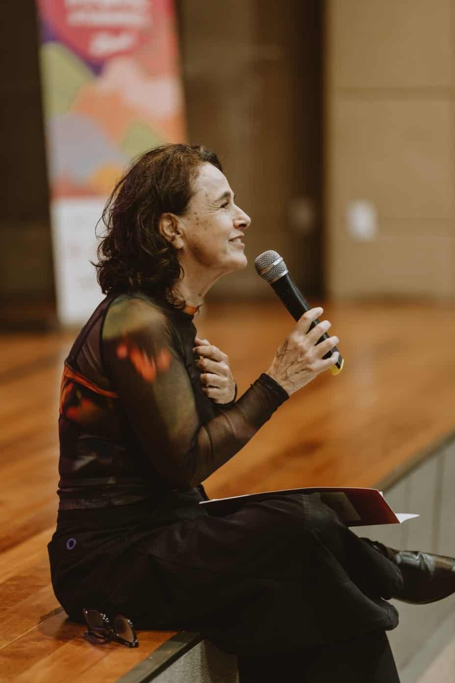 Luiza Lins, diretora da Mostra de Cinema Infantil de Florianópolis, fala ao microfone sentada na borda do palco