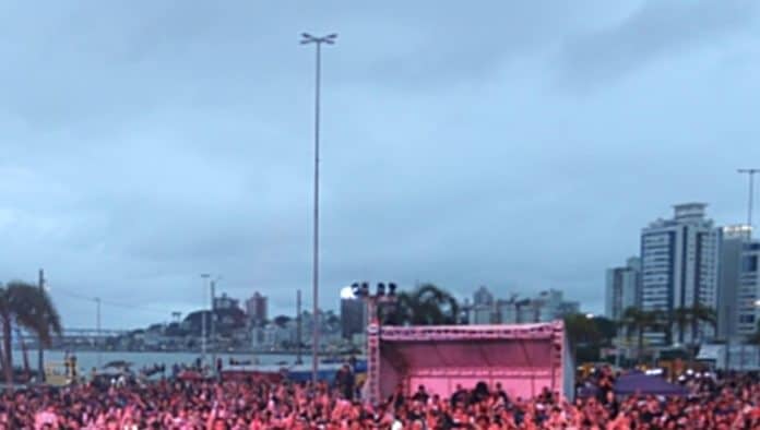 Multidão reunida diante de palco na Beira-Mar Continental, em Florianópolis, com prédios e a baía ao fundo em fim de tarde nublado