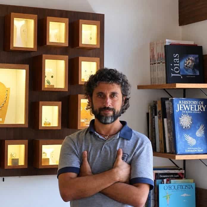 O artista e joalheiro Márcio Sodré, de camisa polo cinza e braços cruzados, em frente a nichos de madeira com joias