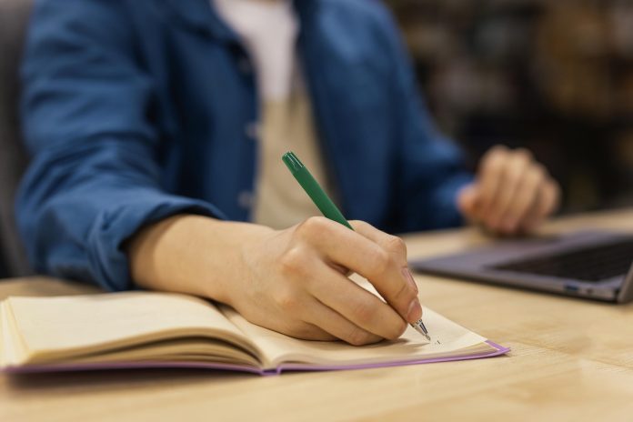 Estudante escreve em um caderno ao lado de um notebook em uma mesa de biblioteca