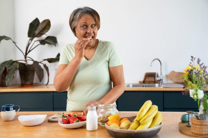Mulher idosa come à mesa da cozinha diante de frutas, como bananas e morangos, e de uma garrafa de leite