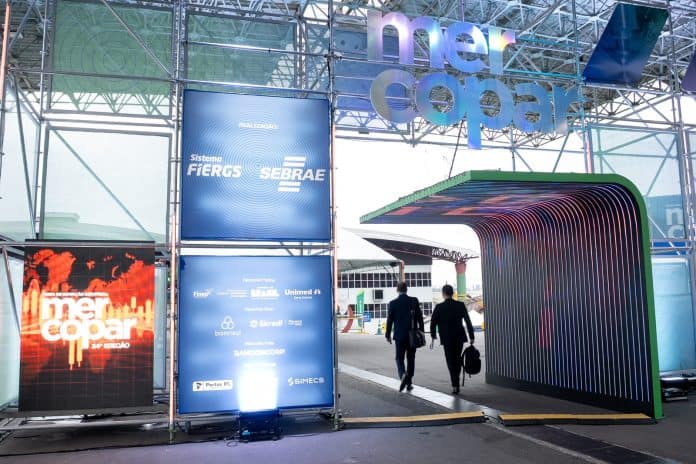 Entrada da Mercopar com letreiro colorido da feira, painéis de patrocinadores e túnel de luz, com dois visitantes de terno caminhando