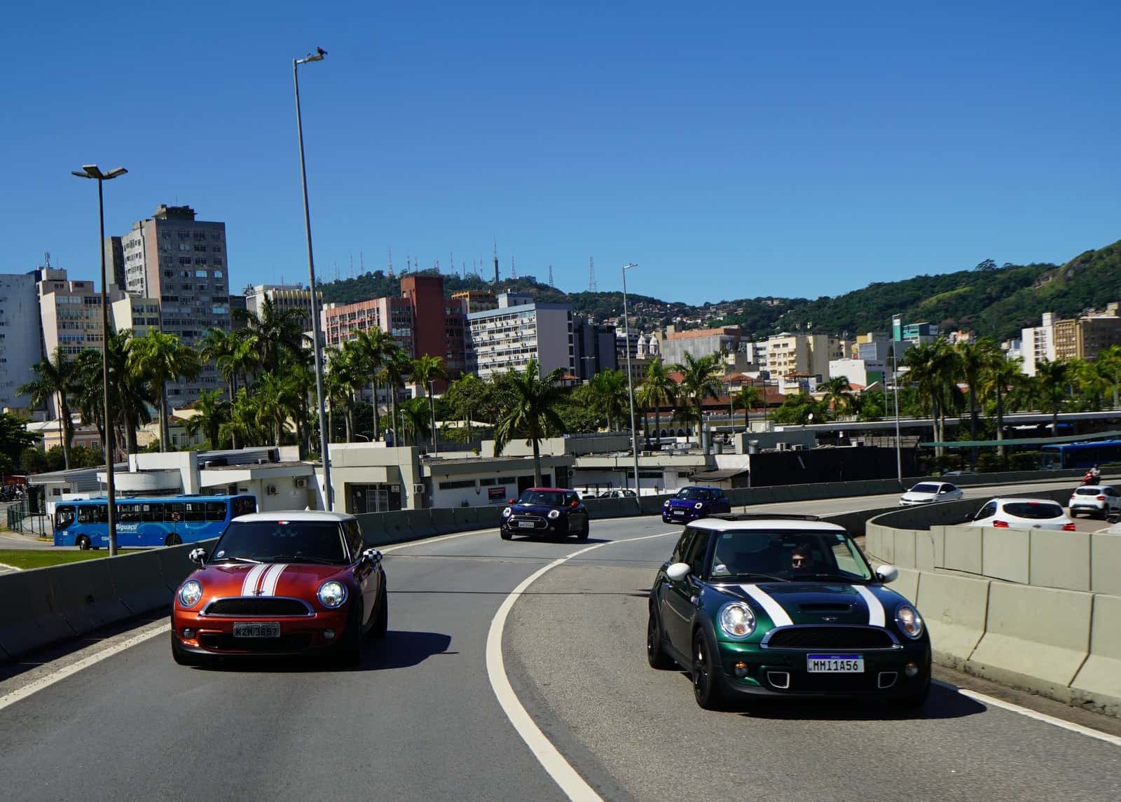 Carros Mini Cooper em comboio por avenida de Florianópolis, com prédios e morros ao fundo