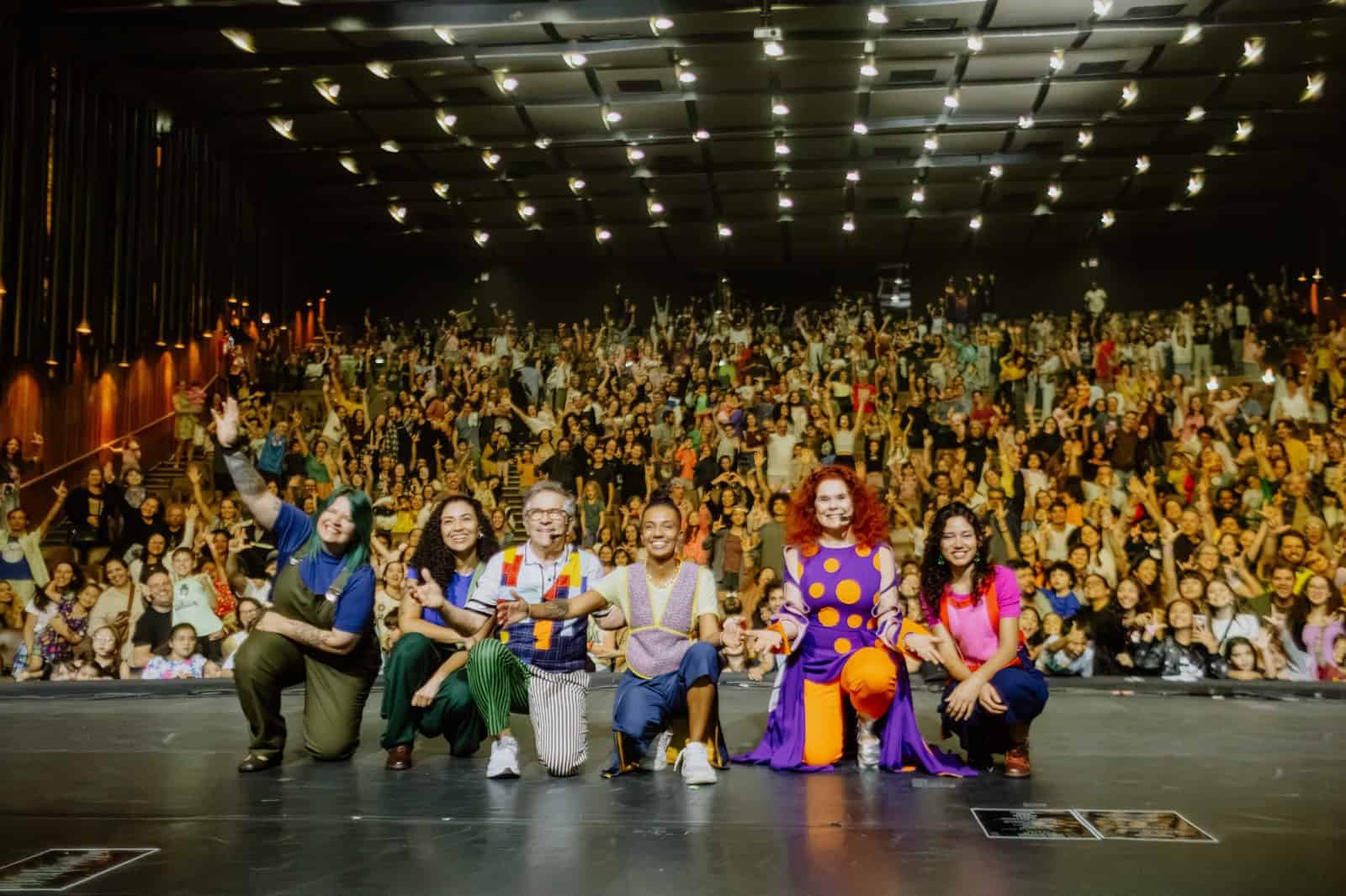 Elenco do show Palavra Cantada 30 anos acena no palco do Teatro Ademir Rosa, com a plateia lotada ao fundo, no encerramento da Mostra de Cinema Infantil
