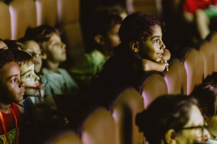Crianças sentadas nas poltronas da sala de cinema do CIC assistindo a um filme da Mostra de Cinema Infantil, com o rosto iluminado pela tela