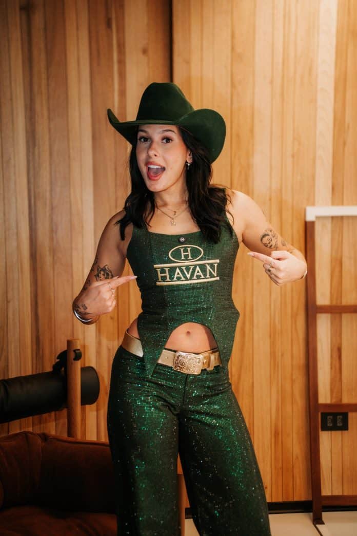 Cantora Ana Castela sorrindo e apontando para o próprio top verde com o logotipo da Havan, de chapéu de caubói verde, calça verde brilhante e cinto com fivela dourada, diante de parede de madeira