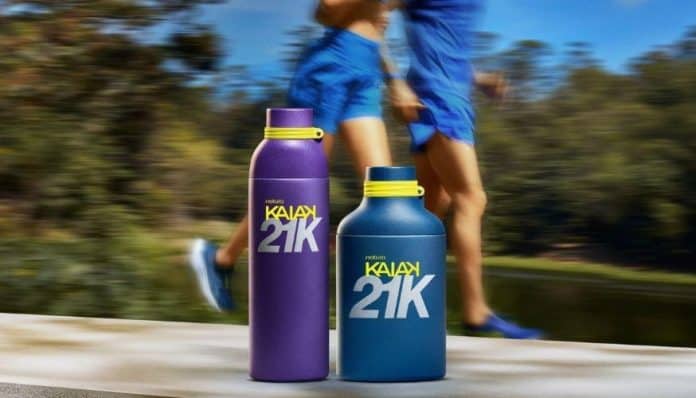 Dois frascos do perfume Natura Kaiak 21K, um roxo e um azul com tampa amarela, sobre uma mureta, com corredores de roupa azul passando desfocados ao fundo em meio à vegetação