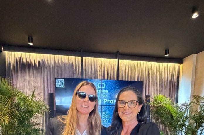 Duas mulheres sorriem lado a lado em ambiente da CASACOR SC, diante de telão com a marca NextMind e a apresentação da palestra