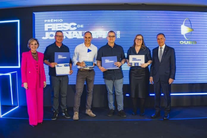 Seis pessoas posam no palco do Prêmio FIESC de Jornalismo 2026, quatro delas segurando certificados, diante de telão com a palavra Ouro