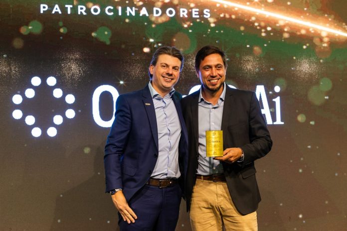 Dois homens sorriem no palco do TOP de Marketing e Vendas ADVB/SC; um deles segura a placa dourada de patrocinadora da OperOS AI, com o painel Patrocinadores ao fundo