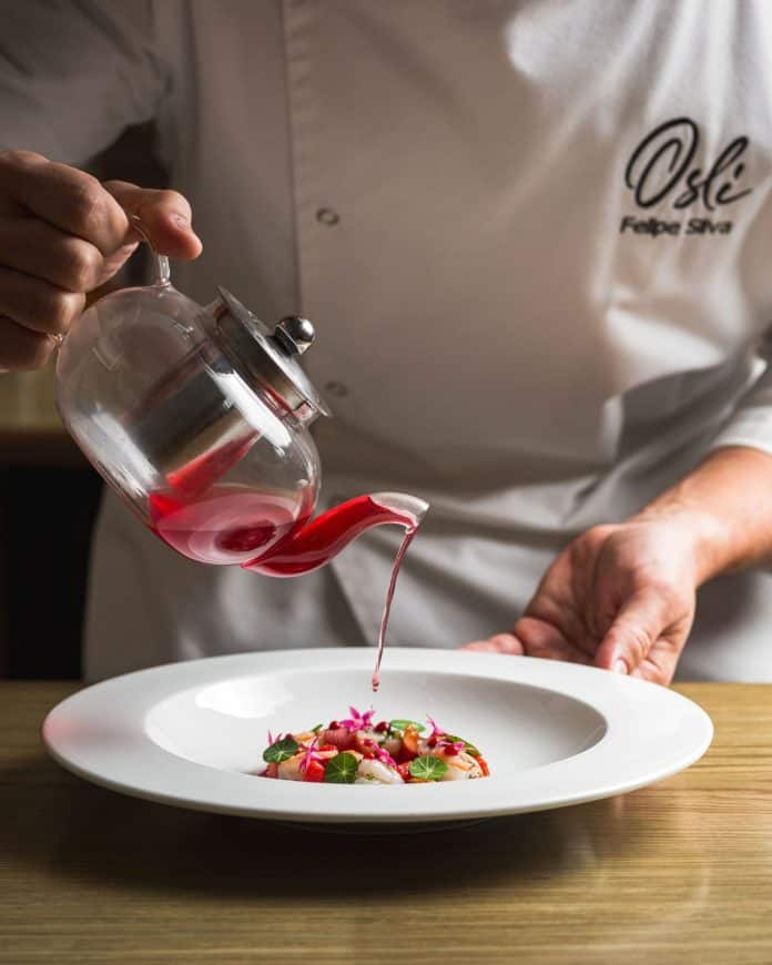 Chef do Osli Restaurante serve caldo vermelho de bule de vidro sobre prato de camarão com flores e capuchinha