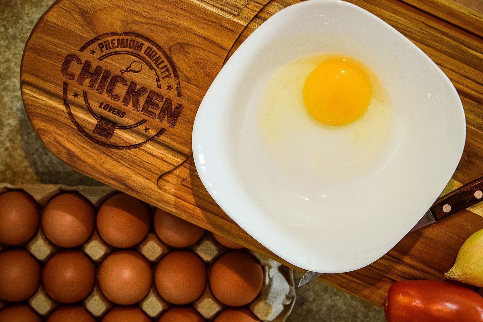Ovo cru em prato branco sobre tábua de madeira, ao lado de uma bandeja com ovos
