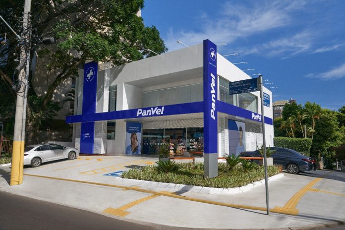 Fachada de loja da Panvel Farmácias, em prédio branco com faixas azuis e o logotipo da rede, com estacionamento e jardim à frente sob céu azul