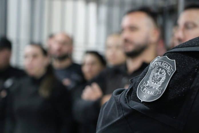 Distintivo da Polícia Penal de Santa Catarina no uniforme de um policial penal, com colegas desfocados ao fundo