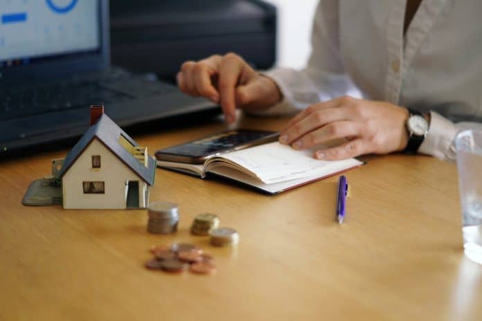 Mãos fazendo contas com calculadora e caderno ao lado de miniatura de casa e moedas sobre mesa de madeira