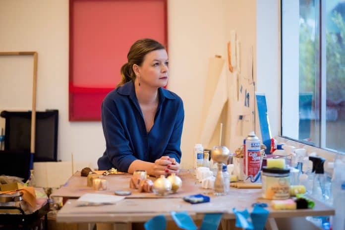 A artista Roberta Tassinari em seu estúdio, sentada à mesa de trabalho com materiais de pintura