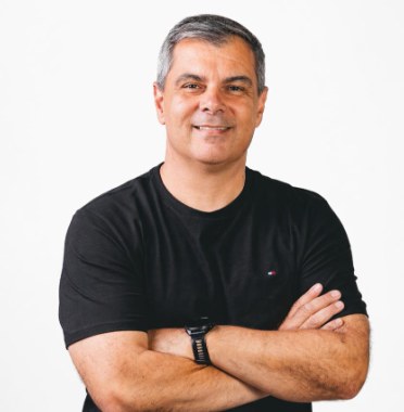 Retrato do comentarista esportivo Rodrigo Faraco, de camiseta preta e braços cruzados, sorrindo diante de fundo branco