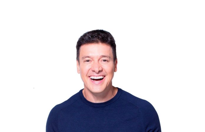 Apresentador Rodrigo Faro sorrindo, de suéter azul-marinho, em foto de estúdio com fundo branco para a campanha do San Satri Hospital Veterinário