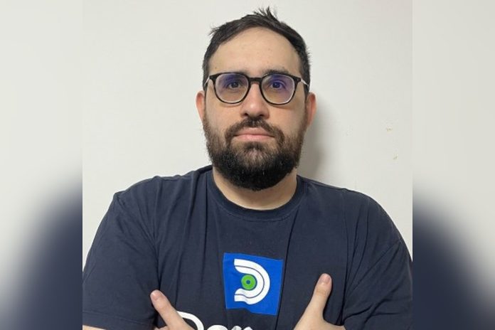 Homem de barba e óculos, de braços cruzados, vestindo camiseta azul-marinho com o logotipo da Deconve