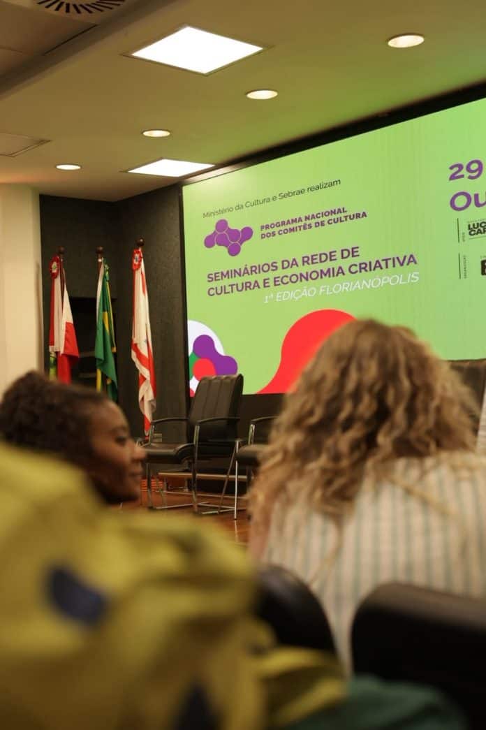 Participantes de costas observam o telão com a identidade visual dos Seminários da Rede de Cultura e Economia Criativa, 1ª edição Florianópolis, no plenário da Alesc