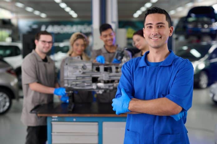 Jovem de macacão azul e luvas azuis sorri de braços cruzados em laboratório automotivo; ao fundo, quatro estudantes de luvas trabalham em uma peça de câmbio sobre a bancada