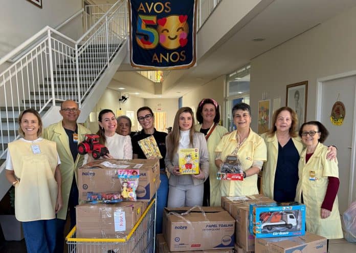 Equipe de gestão do Square SC e diretoria da AVOS com brinquedos e roupas doados, sob banner dos 50 anos da AVOS, em Florianópolis