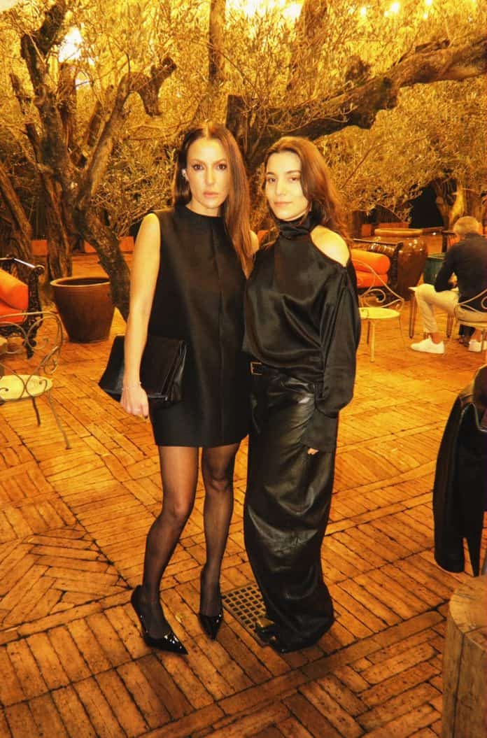 As empreendedoras Victoria Lopes e Paula Freitas, fundadoras da Table For, vestidas de preto em um pátio com oliveiras