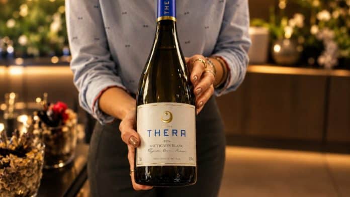 Mão segura garrafa do vinho Thera Sauvignon Blanc 2026, da Vinícola Thera, com rótulo branco e cápsula azul