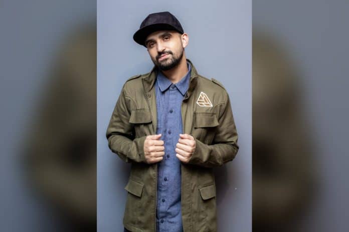 Humorista Thiago Ventura de boné preto e jaqueta verde-oliva posa em fundo cinza-azulado