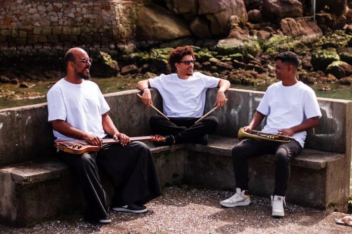 Os três músicos do trio MABI, de camiseta branca, sentados em banco de pedra à beira-mar com baixo, baquetas e trompete