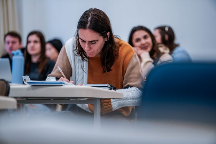 Estudante universitária escreve em caderno durante aula, com colegas desfocados ao fundo