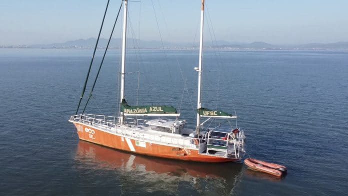 Veleiro ECO, da UFSC, de casco laranja, ancorado no mar calmo com a costa de Florianópolis ao fundo