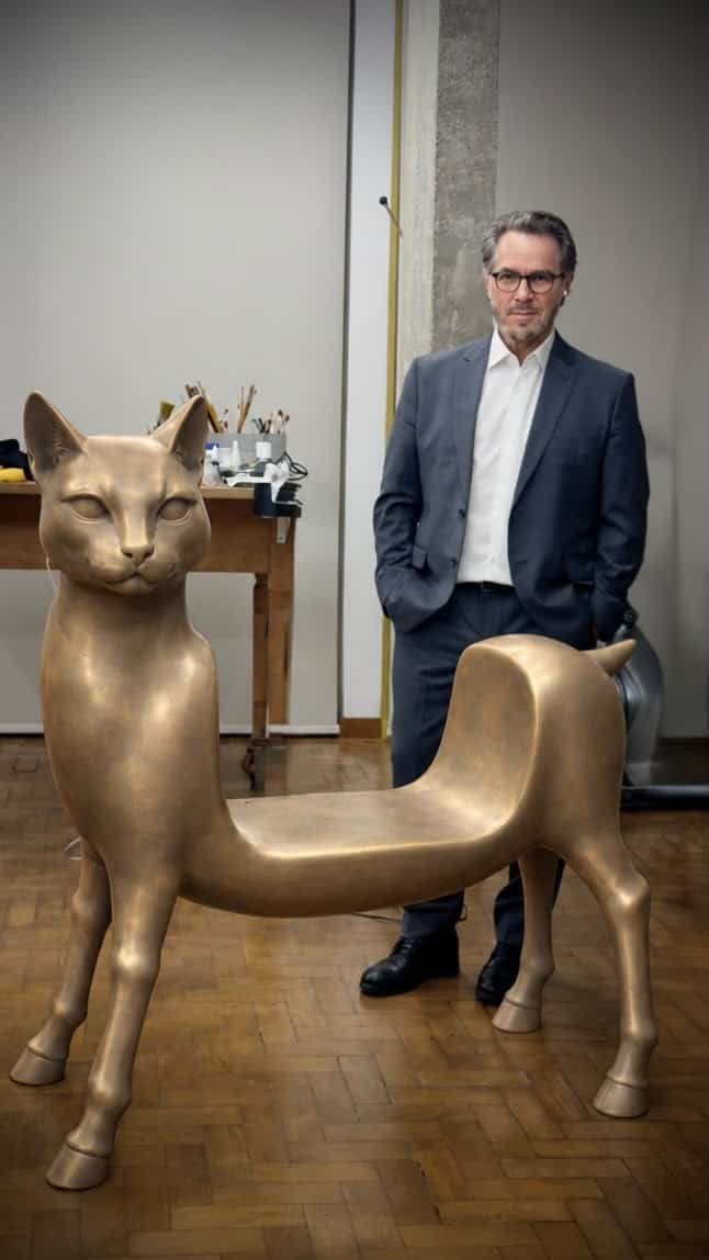 Walmor Corrêa, de terno, ao lado de escultura dourada em forma de banco com cabeça e patas de gato, em ateliê com piso de tacos