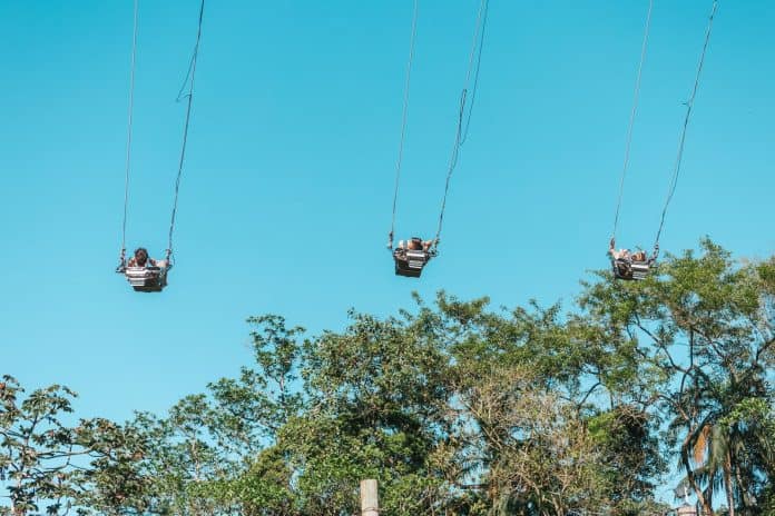 Duas pessoas no balanço Yoop suspensas a grande altura contra o céu azul, com copas de árvores abaixo